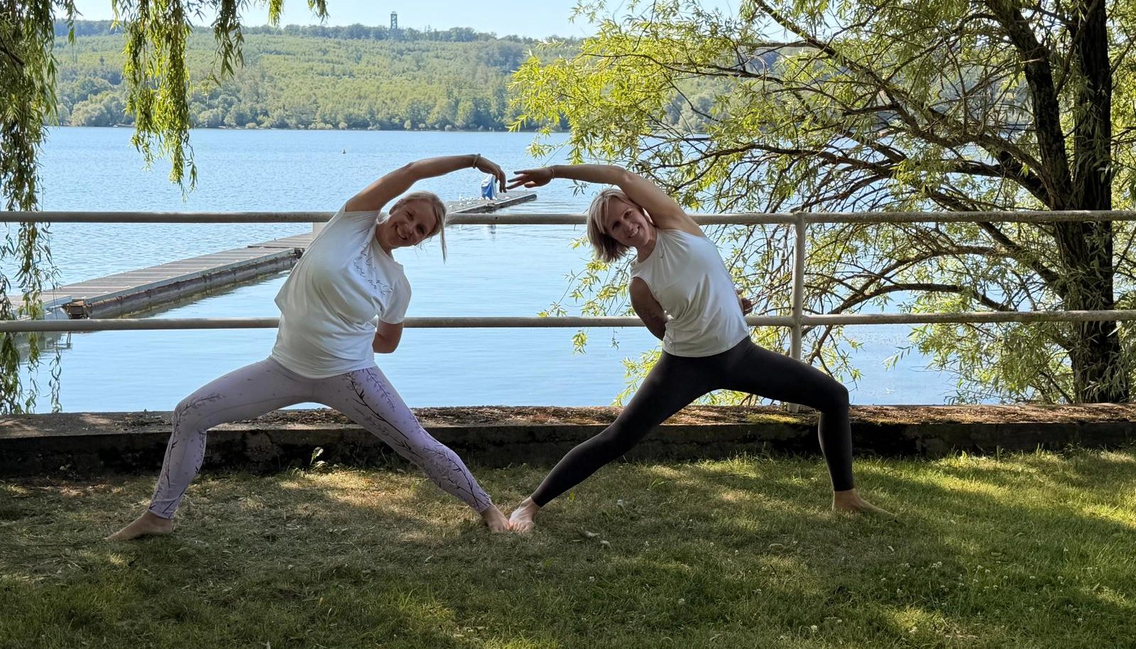 Zwei Frauen üben im Freien eine seitliche Yoga-Haltung auf einer Wiese am See, umrahmt von Bäumen und Steg.
